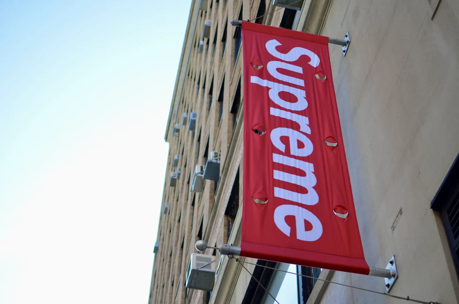 Red Supreme box logo banner hanging from a building facade against a clear blue sky