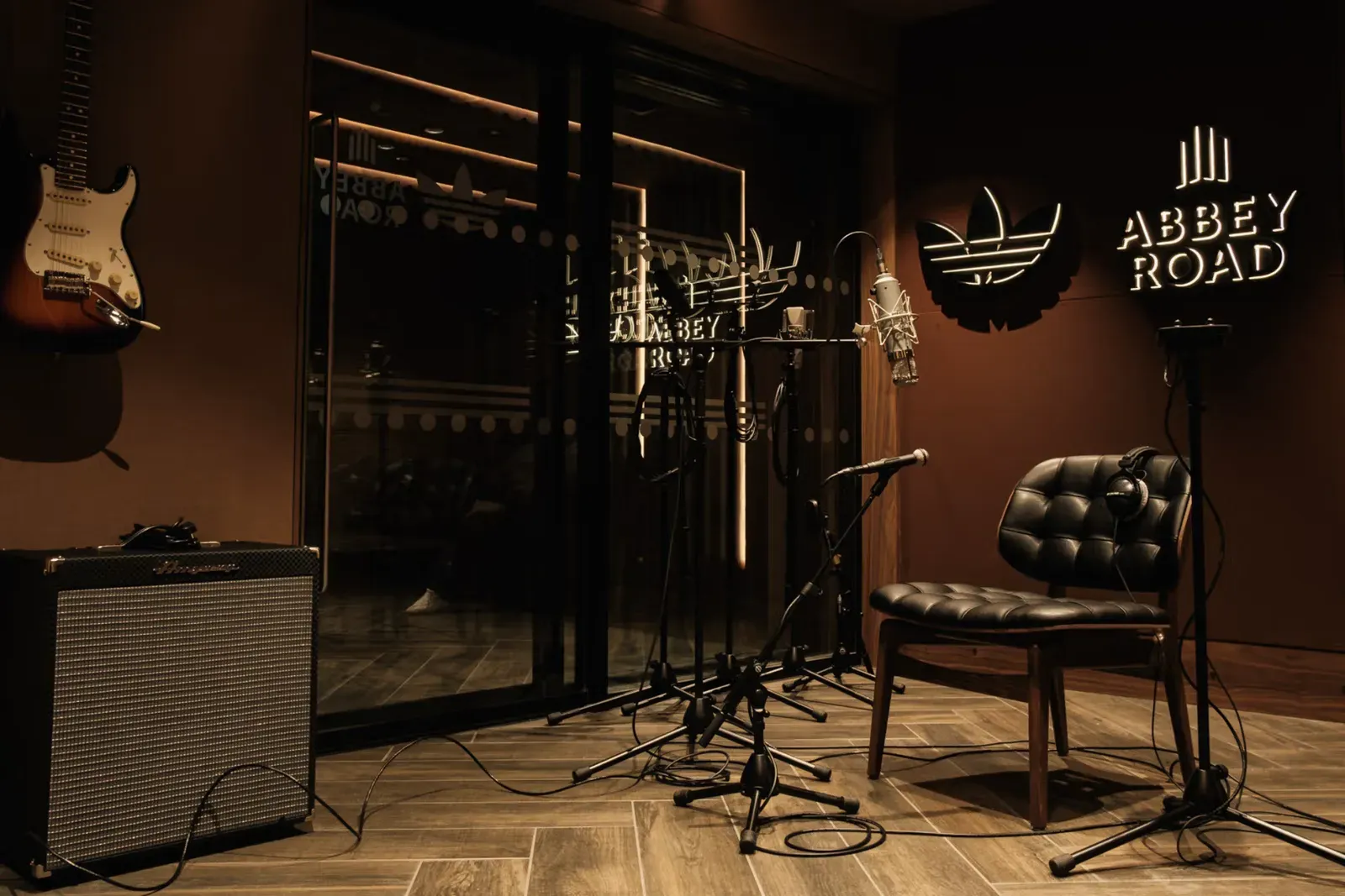 Dimly lit studio corner with mic stands, guitar, amp and glowing Abbey Road and adidas trefoil signs