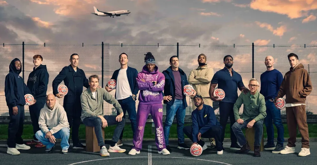 Fourteen men holding footballs pose on a caged pitch at dusk as a plane passes overhead
