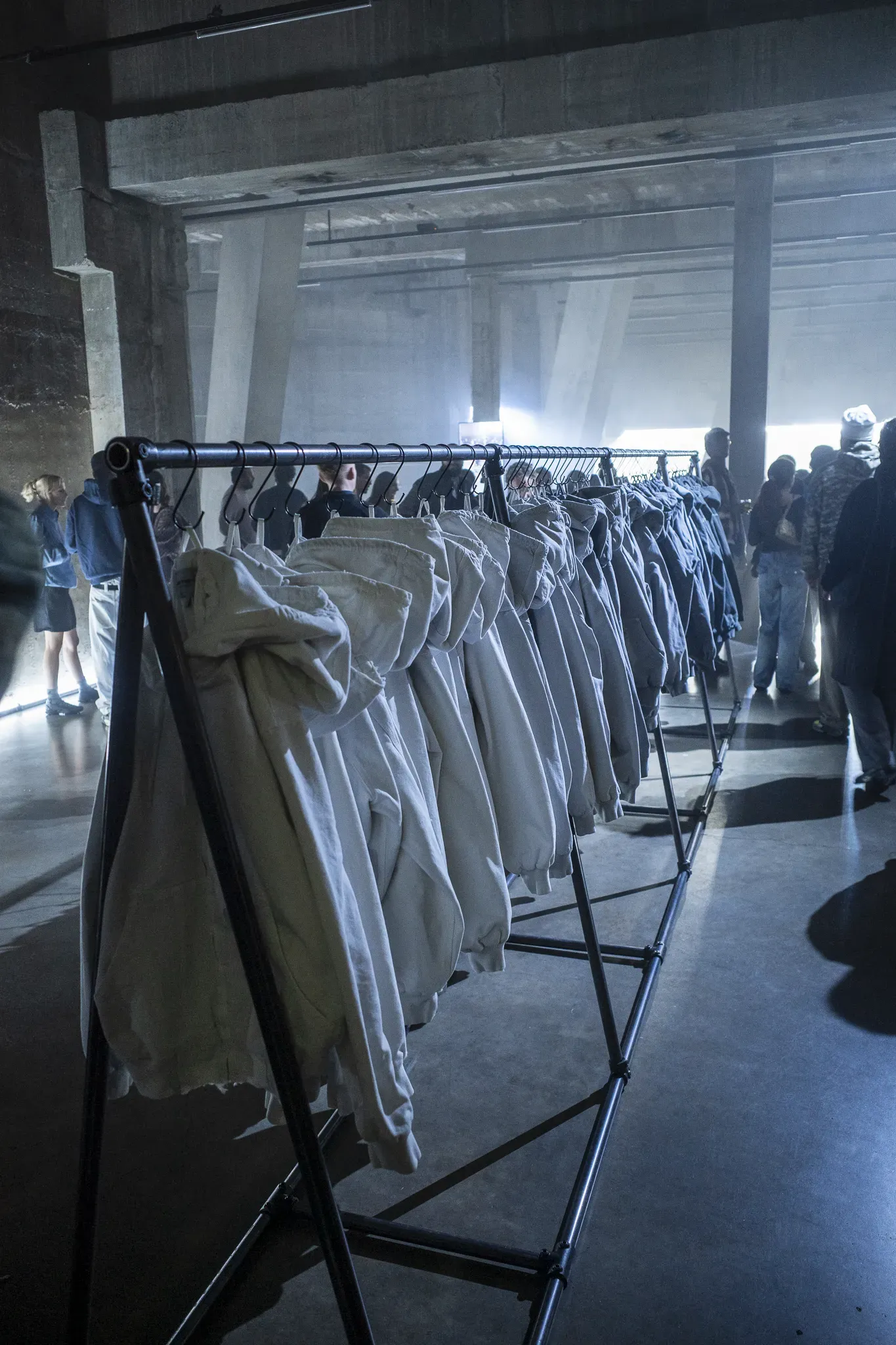 Rail of pale hooded jackets in a raw concrete gallery hall with visitors in the background
