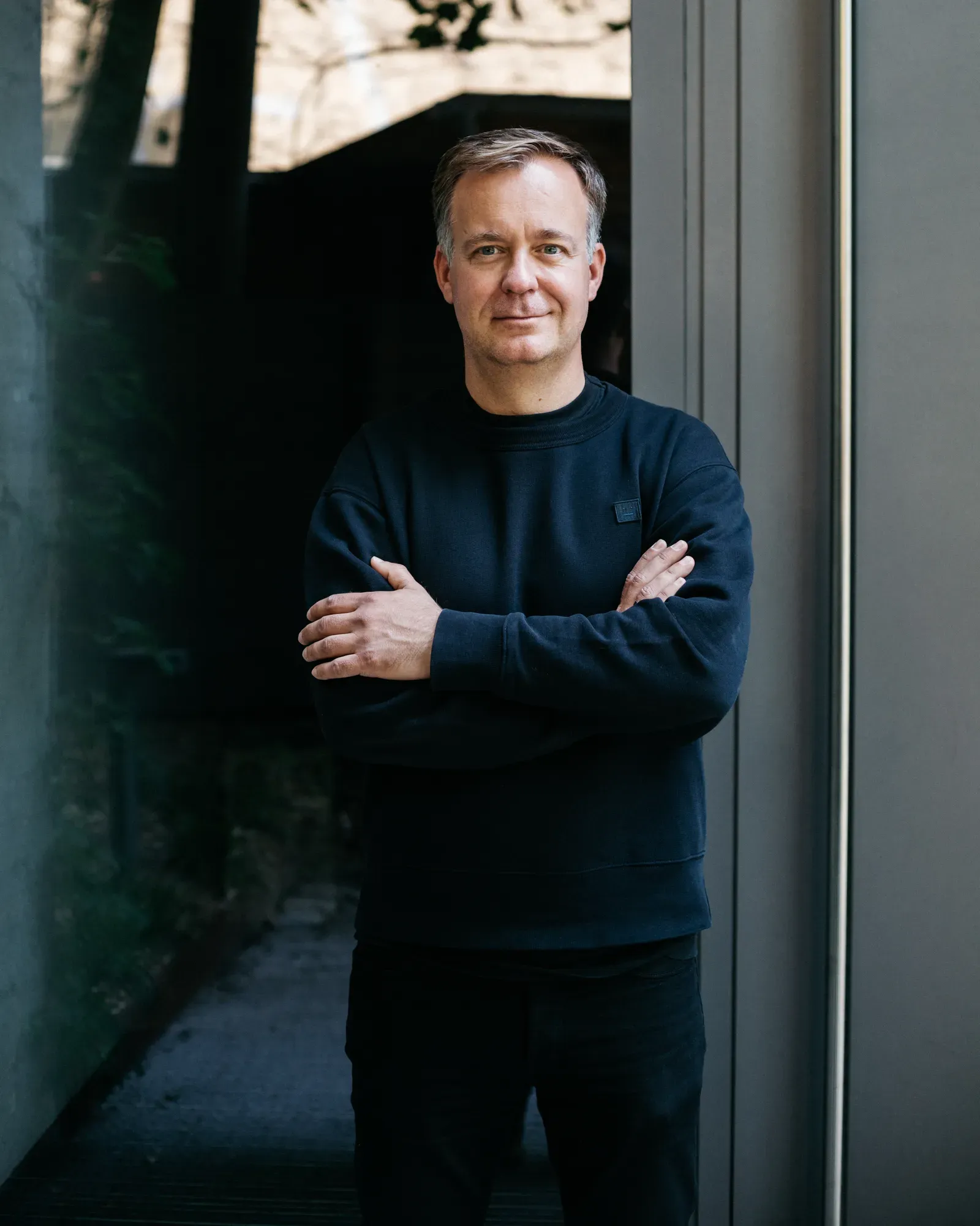 Man in a dark sweatshirt standing with arms crossed in a doorway beside a glass wall
