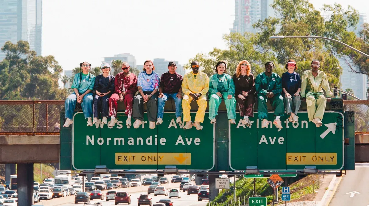 Twelve people in colourful tracksuits sit atop a green freeway sign above heavy Los Angeles traffic