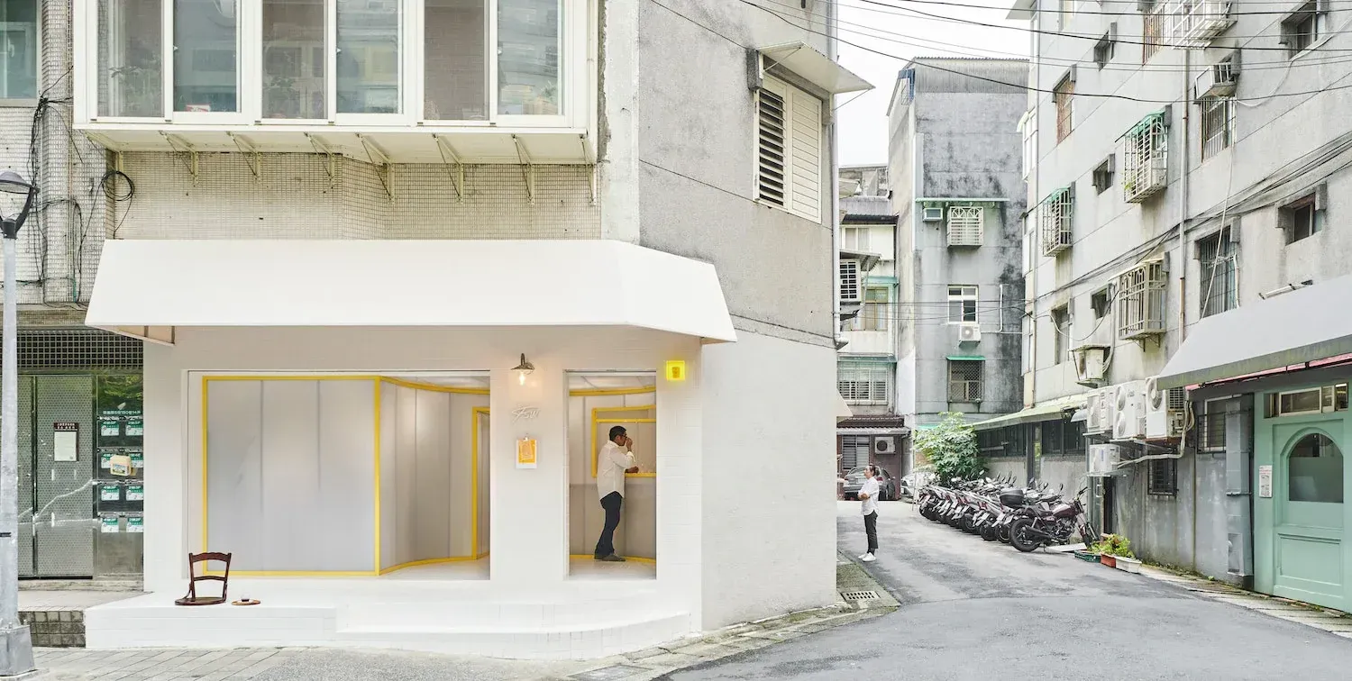 Small white corner shop with yellow window frames on a narrow street lined with scooters