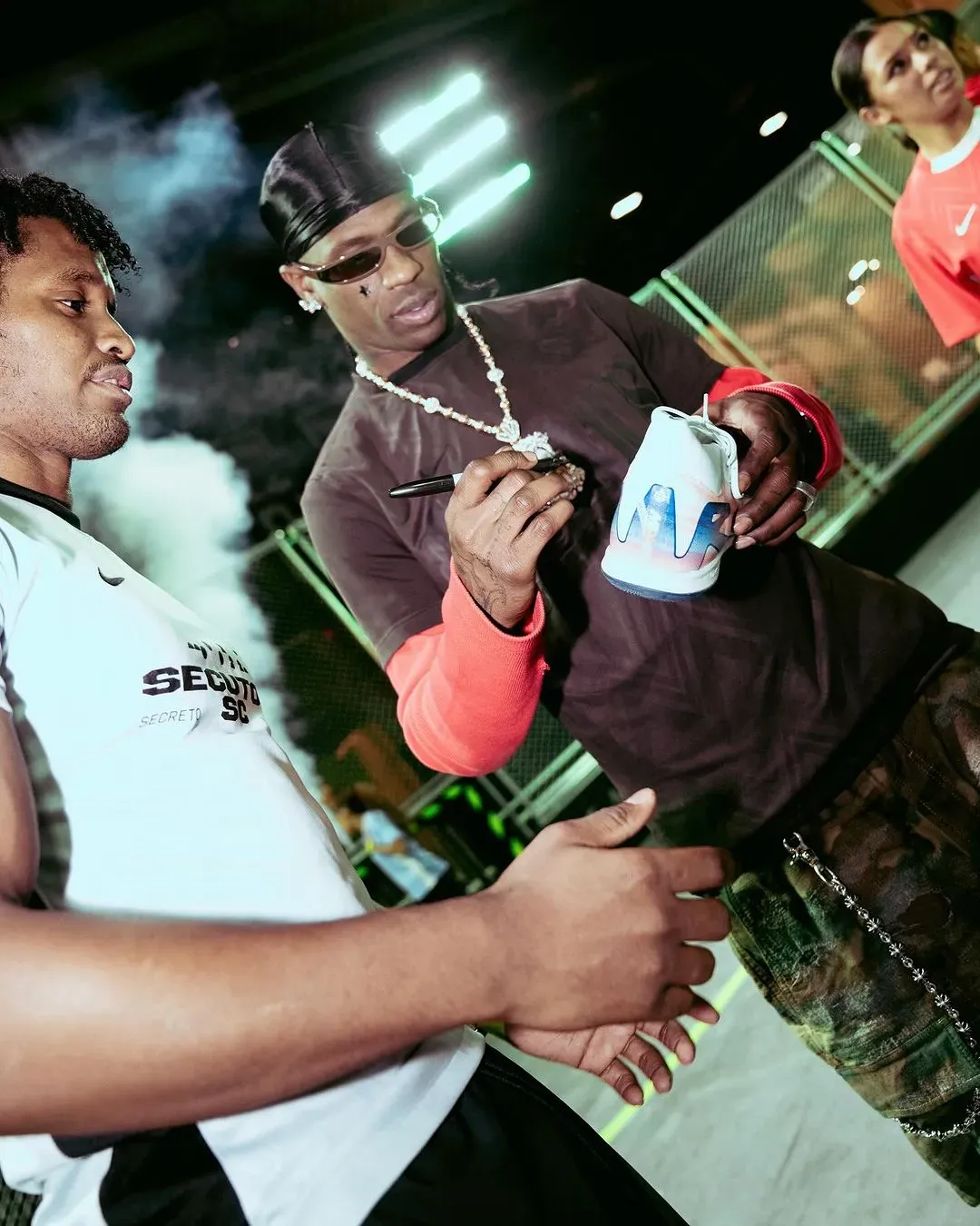 Rapper in durag and sunglasses signing a white Nike sneaker for fans at a neon-lit event