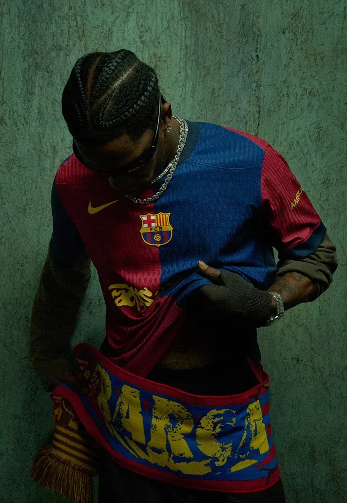Man in braids and sunglasses lifts the hem of the FC Barcelona Cactus Jack jersey, a Barça scarf around his waist