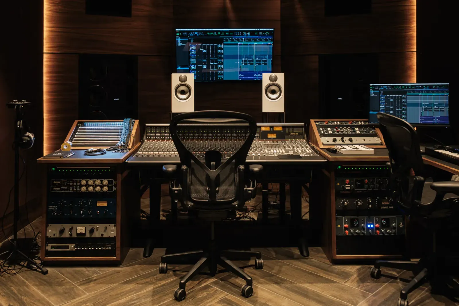 Studio control room with a large mixing console, an office chair, two white monitor speakers and racks of outboard gear