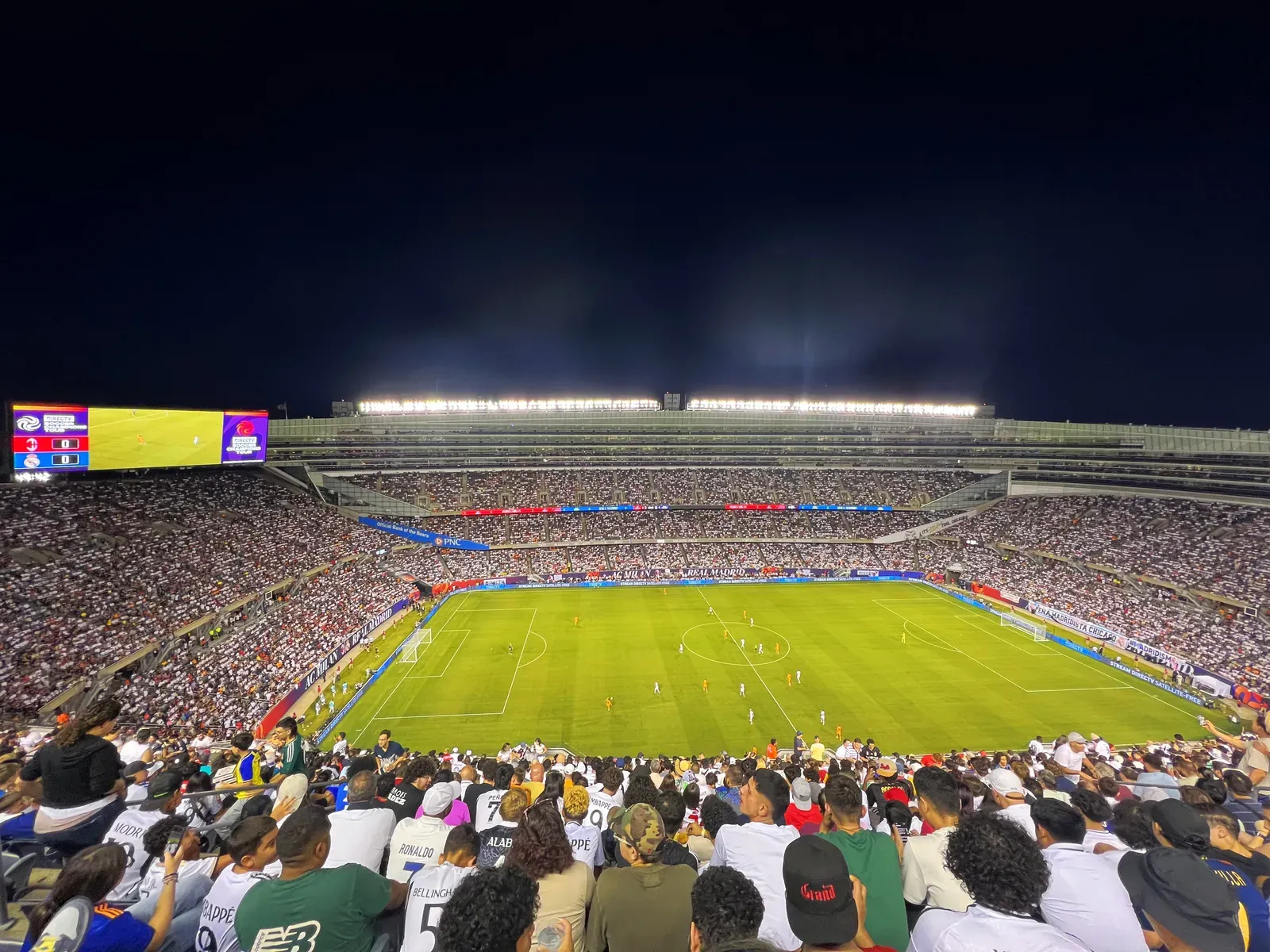 Packed stadium at night as Real Madrid play AC Milan, with fans in white Madrid shirts filling the stands in front
