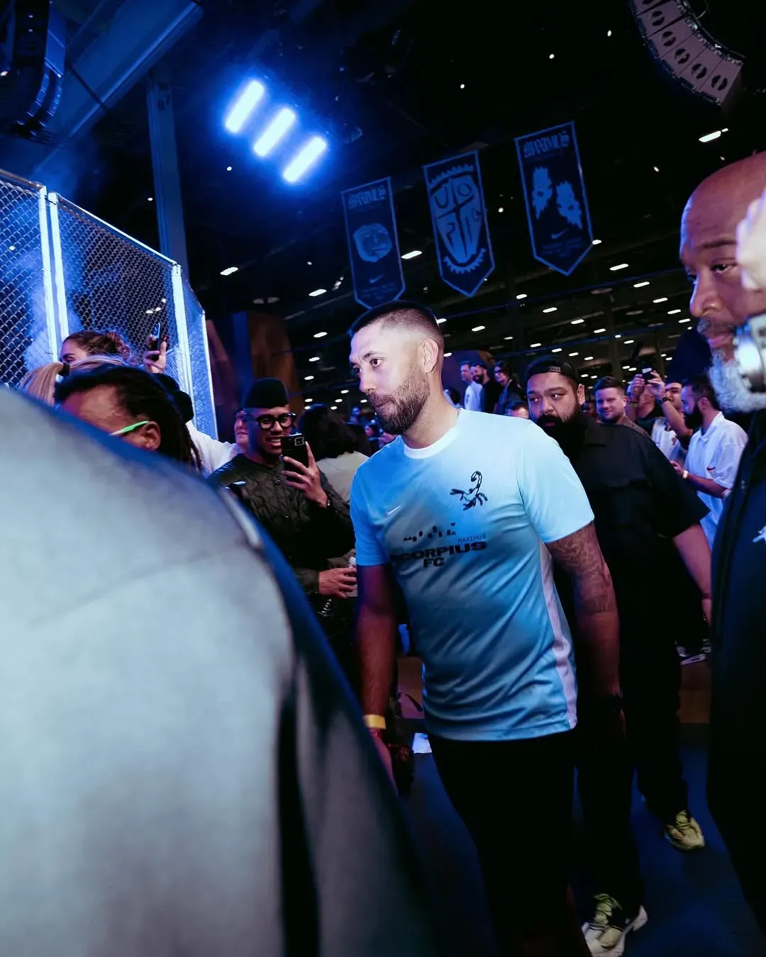 Bearded player in a light blue Scorpius FC jersey walks through a crowd filming on phones beside the caged pitch