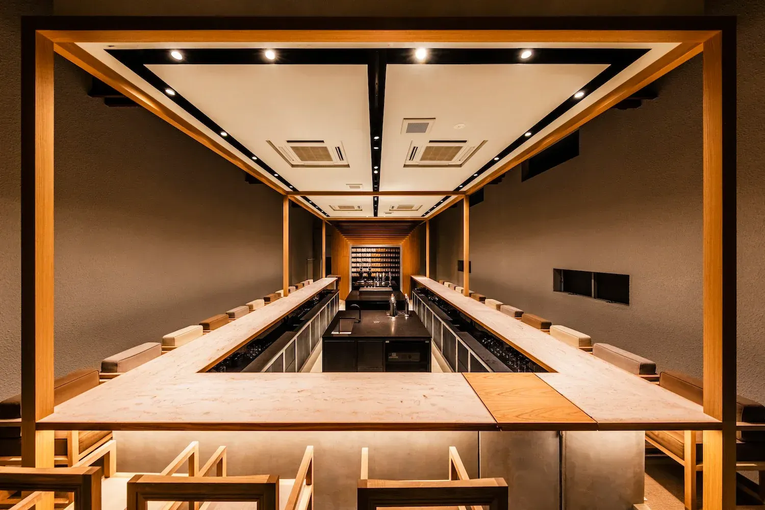 Minimalist café with a long pale-wood counter wrapped around a dark bar, framed by timber posts in warm light