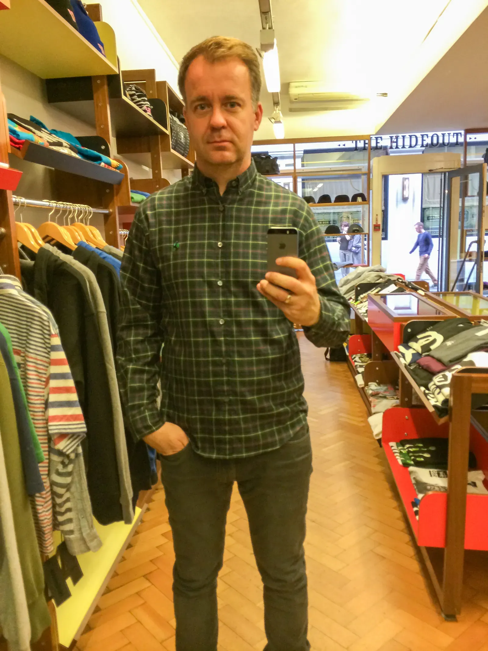 The author tries on a green check shirt in a mirror selfie inside The Hideout, wooden shelves and parquet behind