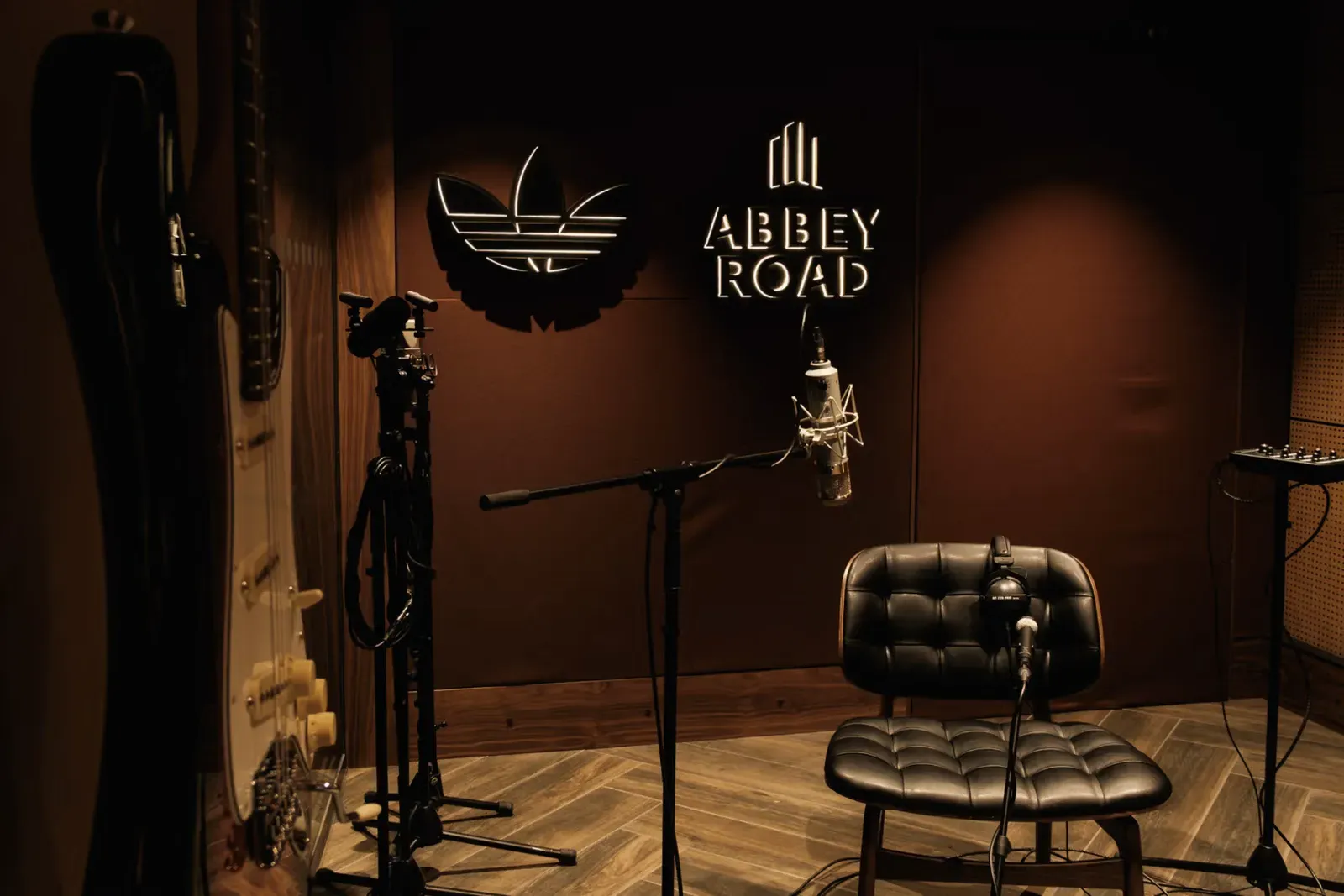 Recording room with a condenser mic, guitars and a leather chair beneath backlit adidas trefoil and Abbey Road signs