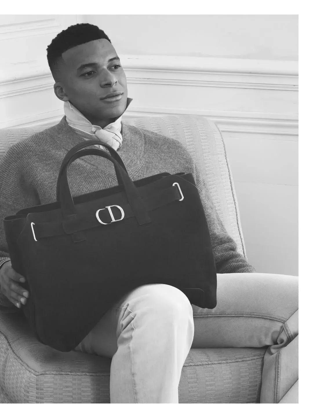 Mbappé in a black and white Dior campaign shot, seated in an armchair with a large dark CD-logo tote on his lap