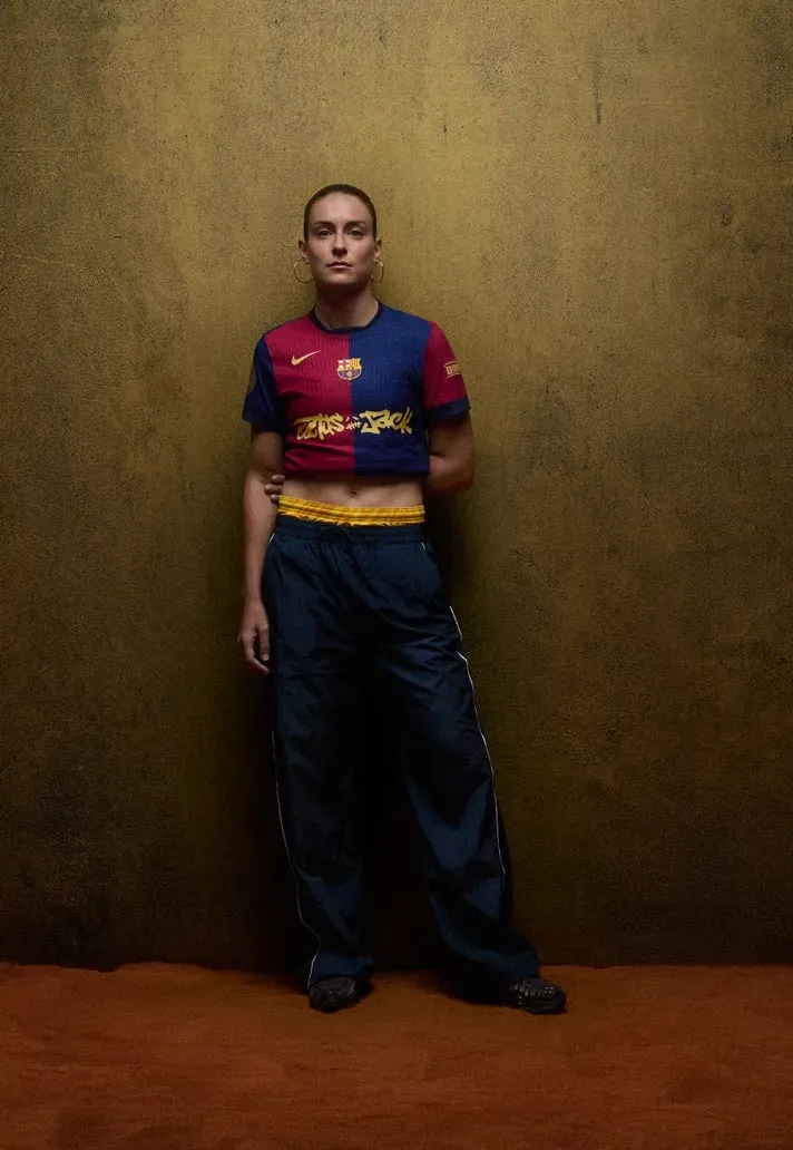 Woman in a cropped FC Barcelona jersey with gold Cactus Jack lettering and navy track pants against an ochre wall