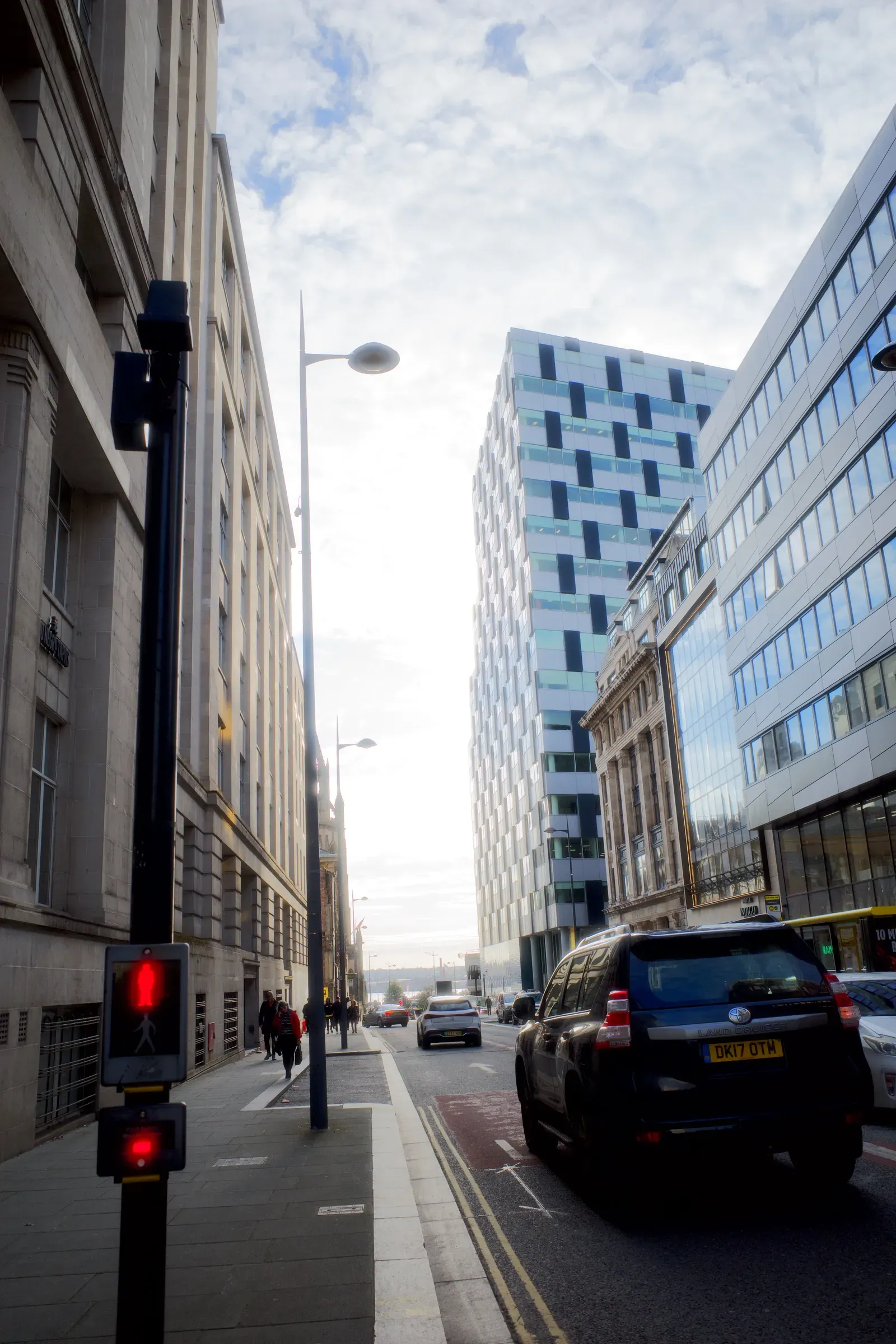 Liverpool city street with a red pedestrian light, stone office blocks and a chequered glass tower under a cloudy sky