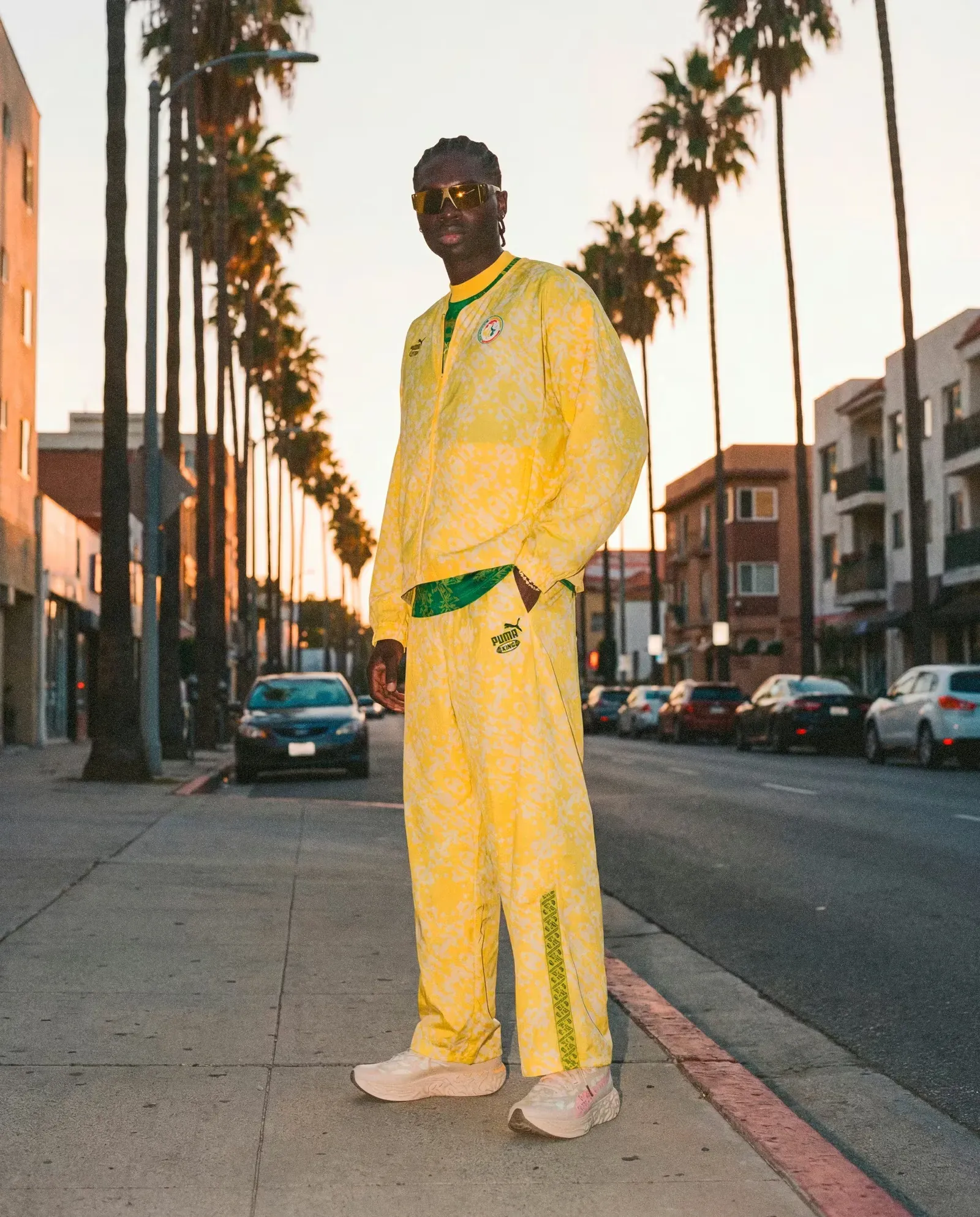 Man in sunglasses and a yellow Puma KING tracksuit with green trim and a team crest on a palm-lined street at sunset