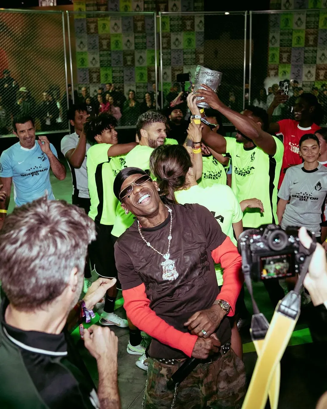 Players in neon Flamma Crew jerseys lift a silver trophy as a laughing man in durag and sunglasses celebrates in front