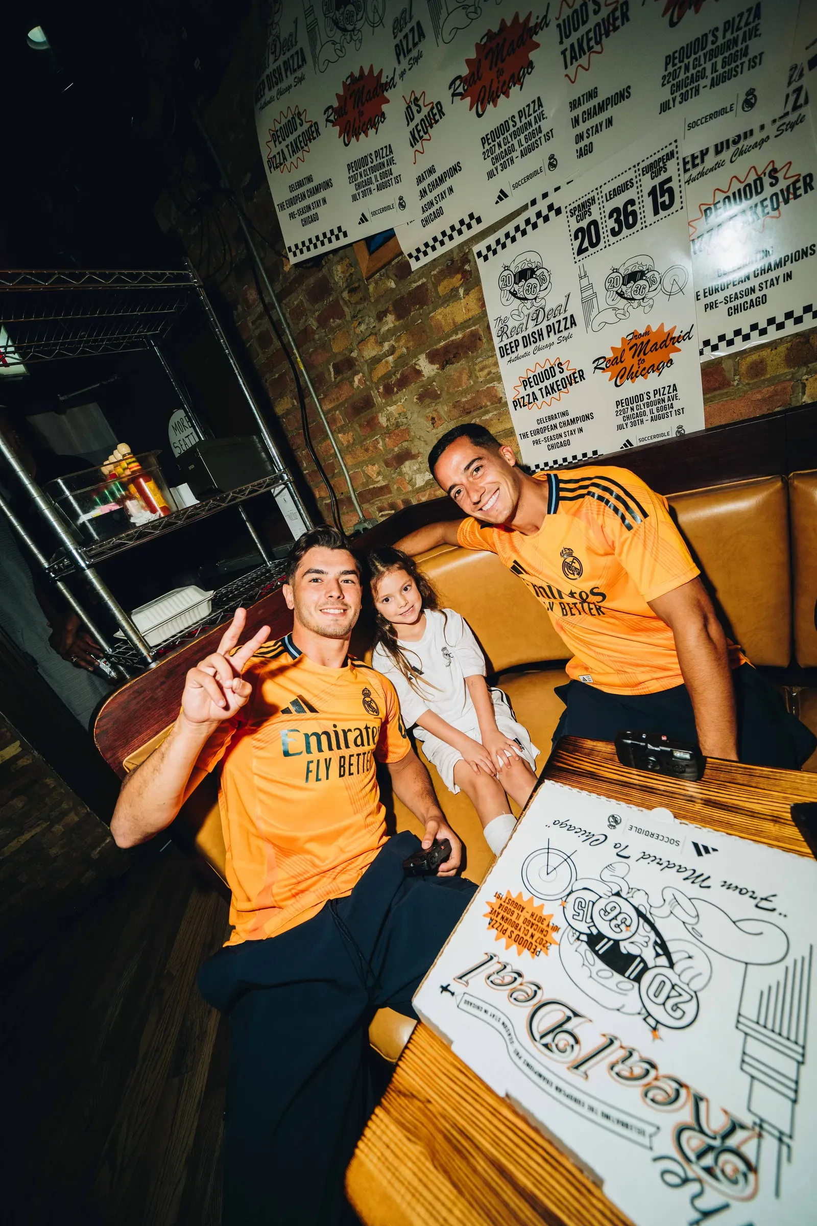 Two men in orange Real Madrid shirts sit with a girl in a white kit under Pequod’s Pizza takeover posters