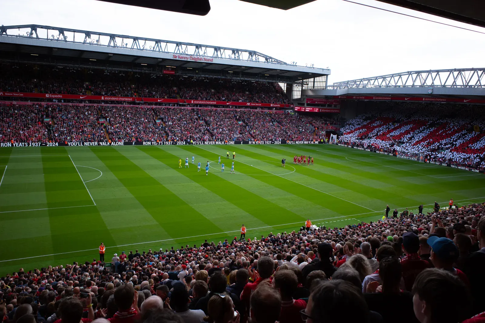 Full Anfield crowd with a red and white card mosaic, boards reading Ron Yeats 1937–2024 and You’ll Never Walk Alone