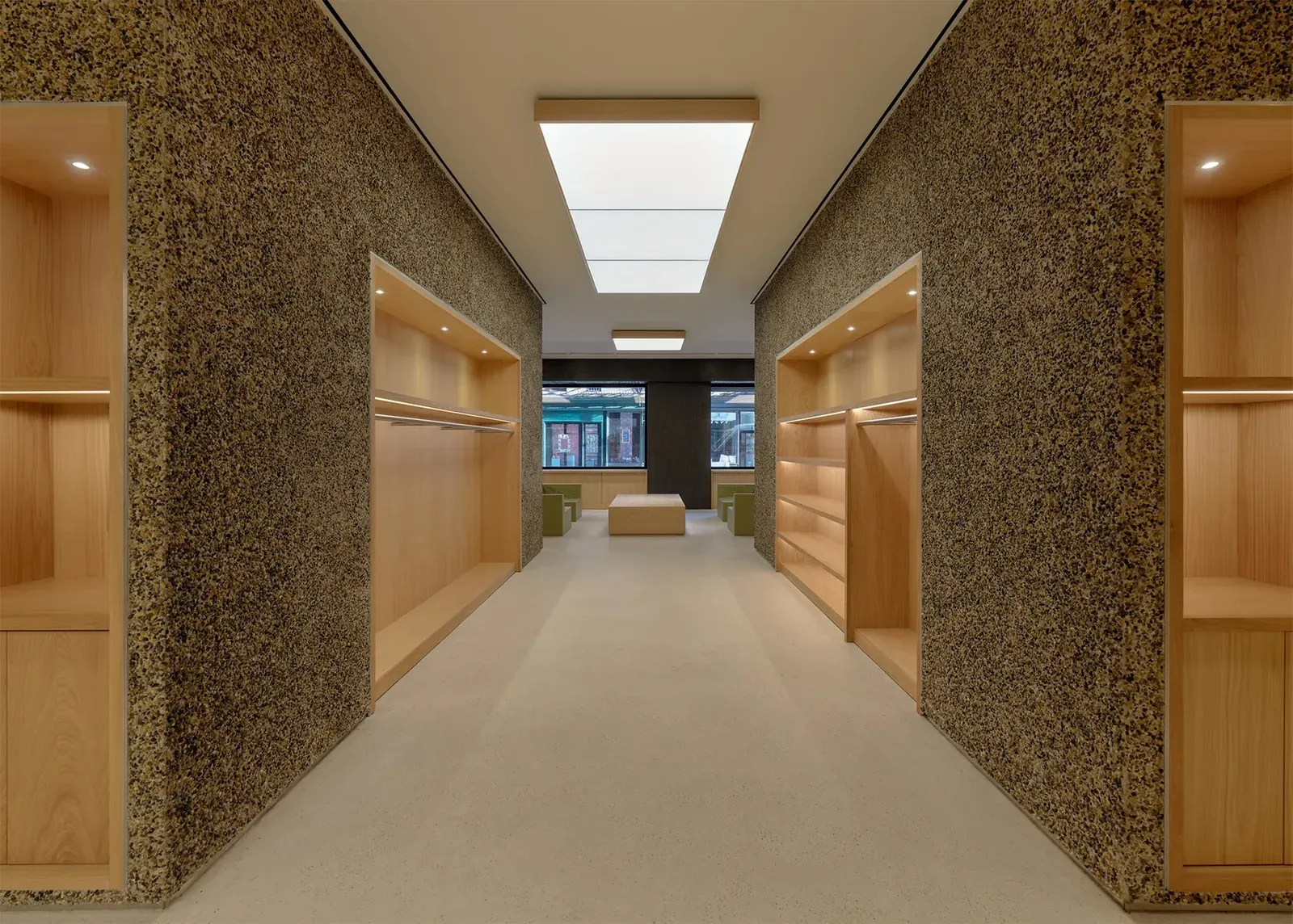 Aisle between pebble-clad walls with empty, lit oak shelf niches, leading to the lounge seats by the street windows