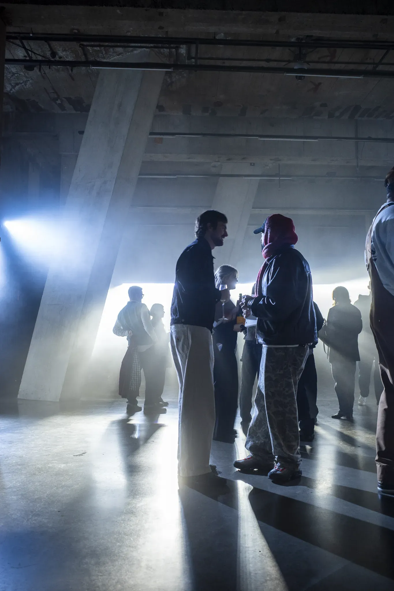 Guests talk in a hazy concrete hall, backlit by bright white light streaming between slanted columns