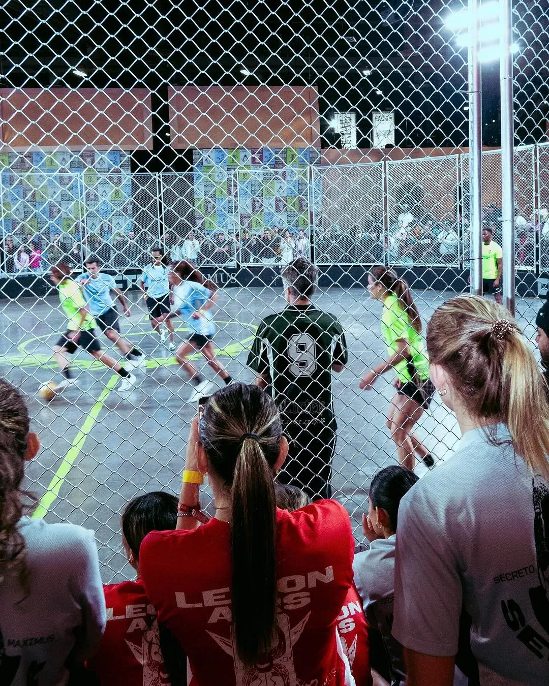 Spectators in red Legion Airs shirts watch through the chain-link cage as neon and light blue teams chase the ball