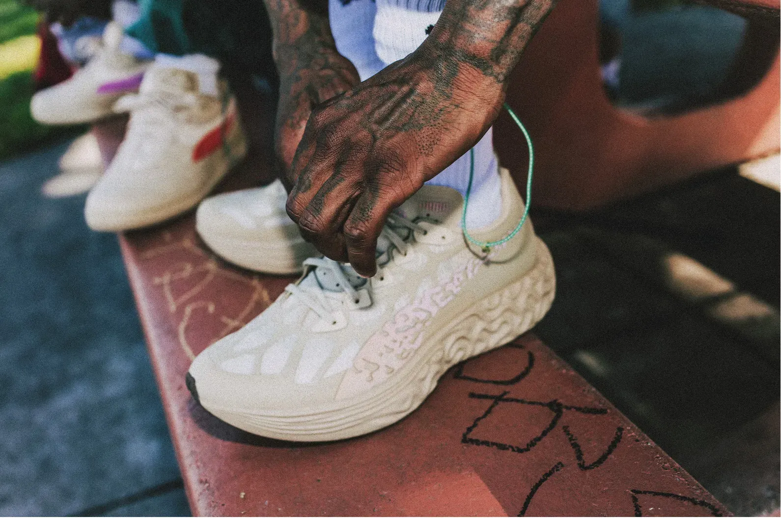 Tattooed hand lacing a cream sneaker with a wavy sculpted sole, the foot propped on a red bench covered in scribbles