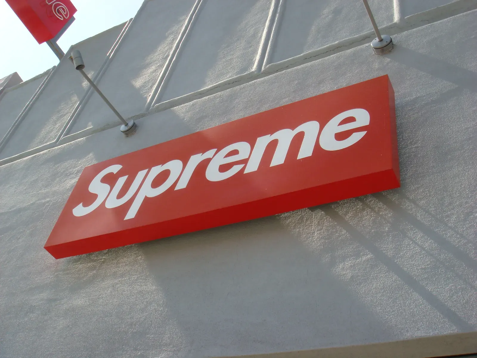 Looking up at the red Supreme box logo sign on the store’s grey stucco facade in bright sun