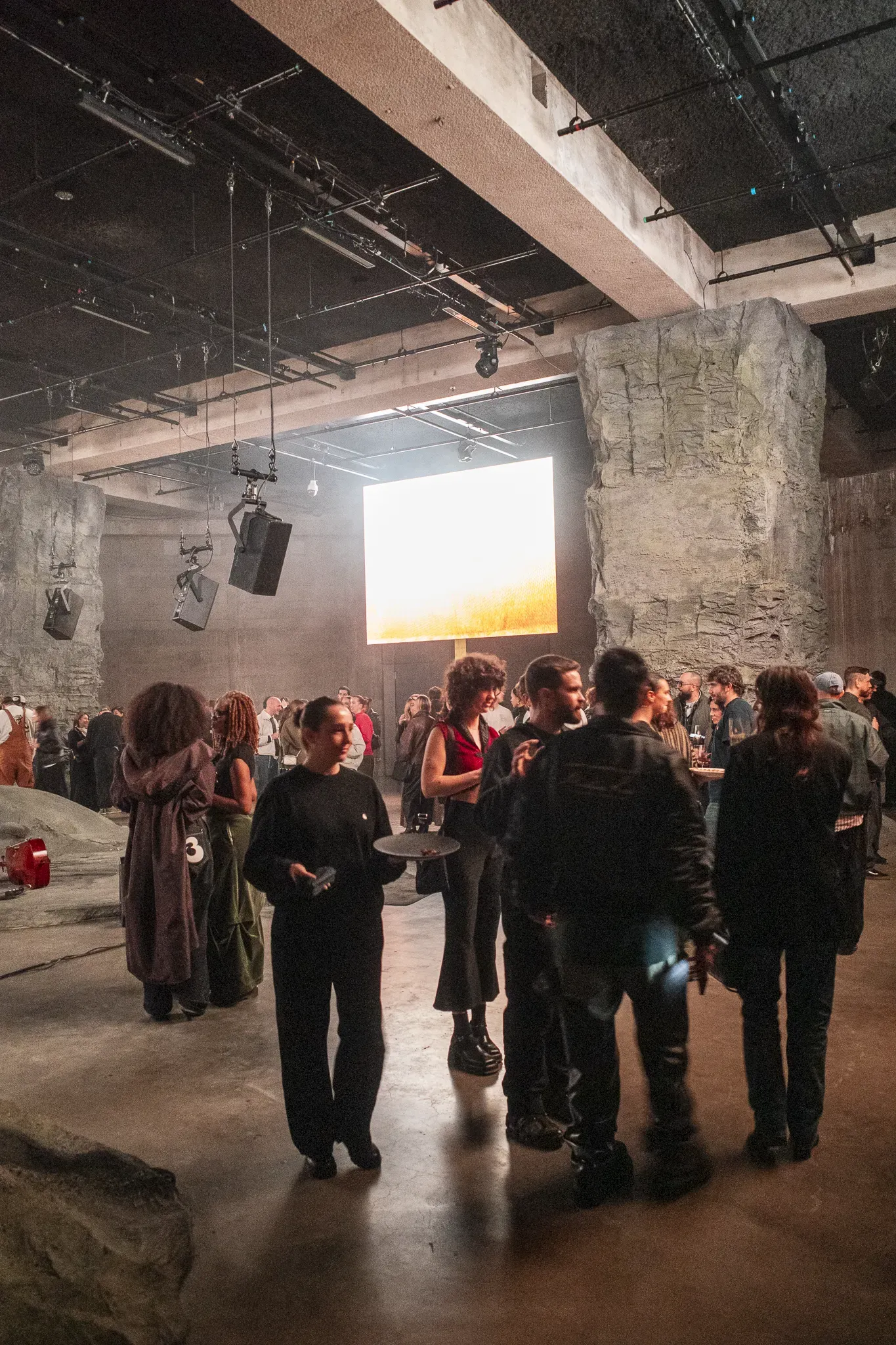 Crowd mingling between rock-like pillars under hanging speakers, a glowing screen with a golden image behind