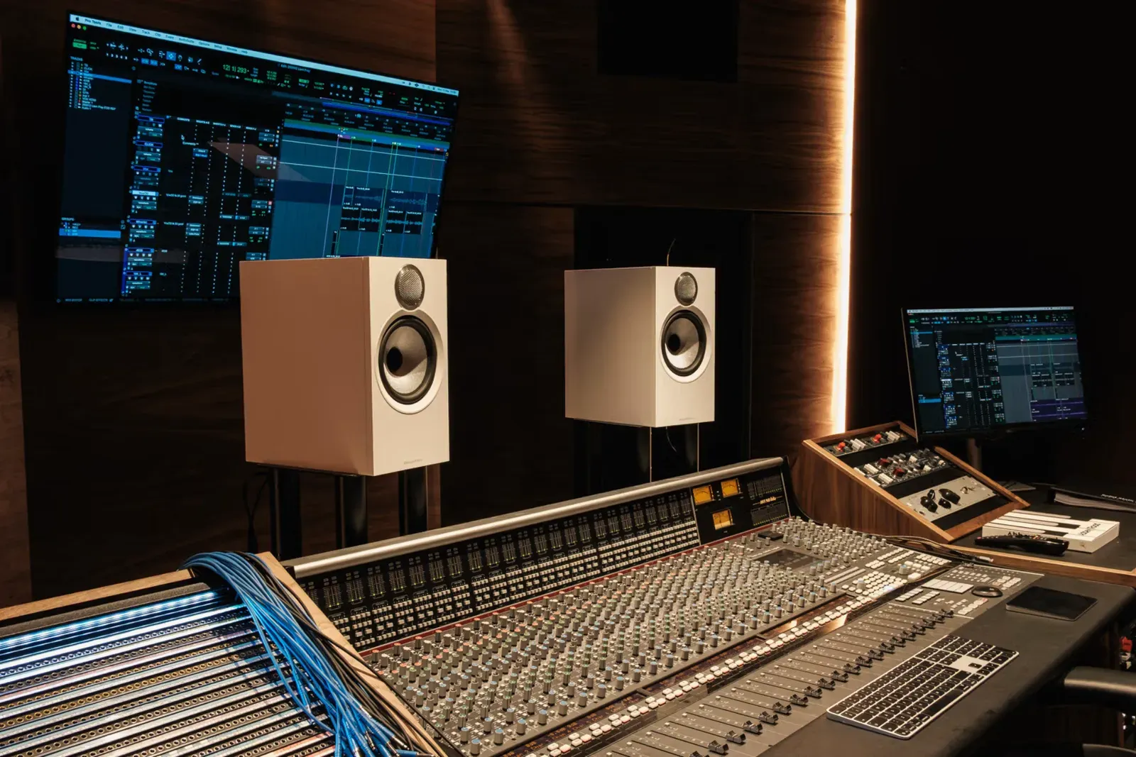Large mixing console with a patch bay of blue cables, white monitor speakers and screens showing a recording session