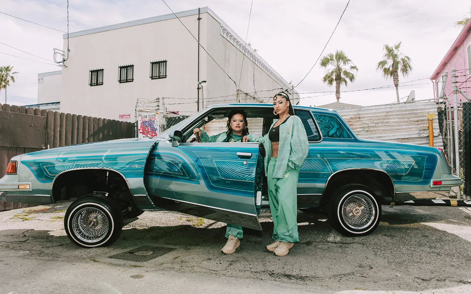 Two women in mint-green Puma tracksuits pose by a teal lowrider with its door open, palm trees behind the fenced lot