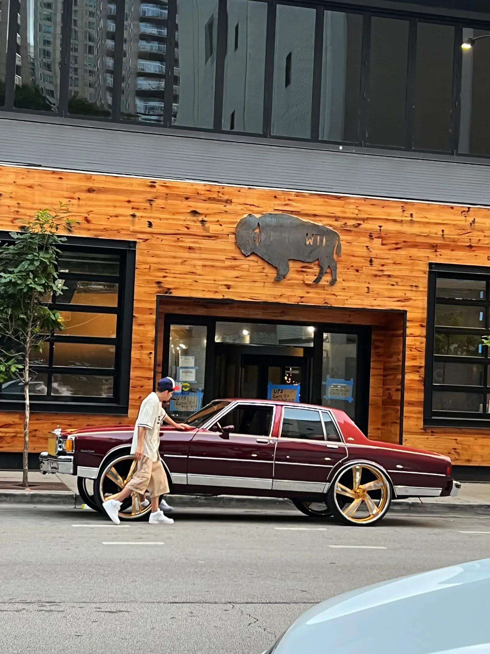Man in a cap running a hand along a maroon vintage sedan on huge gold rims outside a wood-clad front with a bison sign
