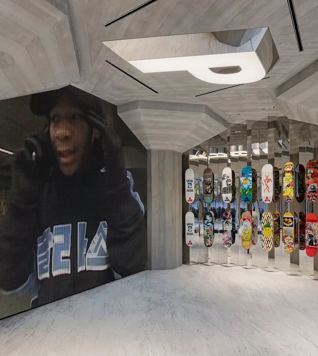 Video wall of a person in a cap and a hoodie with Korean lettering, next to hanging skateboard decks, some printed SEOUL