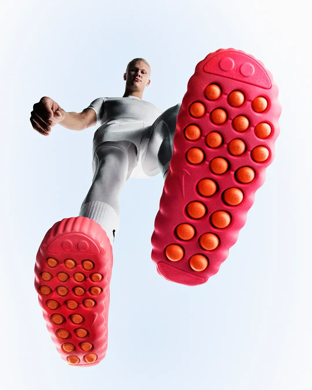 Man in white sportswear shot from below as he strides over the camera, showing the orange foam nodes on both red soles
