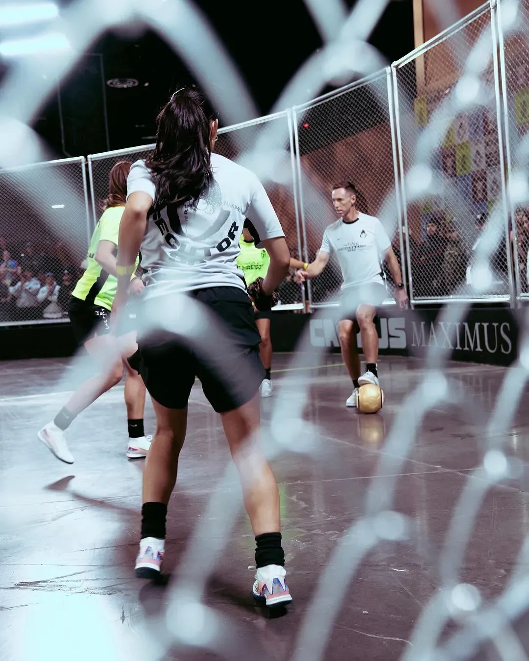 Seen through the blurred cage fence, players in Secutor SC and neon jerseys face off over a gold ball
