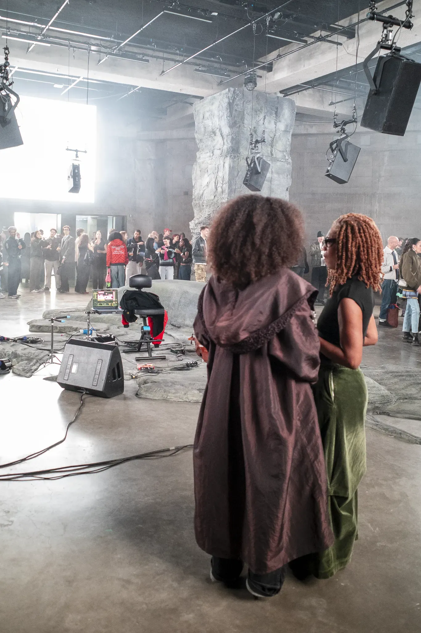 Two guests seen from behind look across a rock-like stage with a stool, laptop and speaker toward the crowd