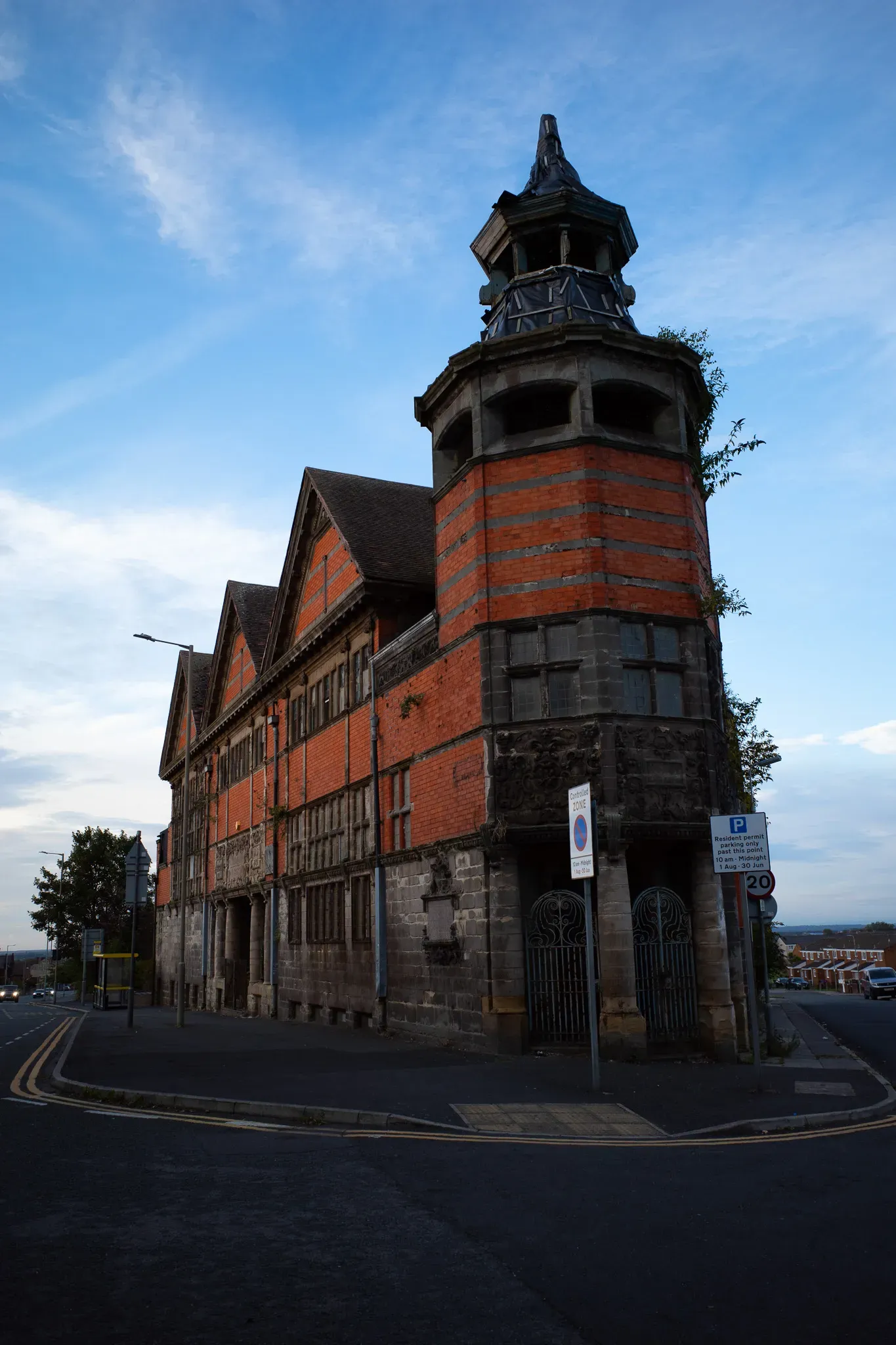 Weathered Victorian building of red brick and stone with an ornate corner tower, plants growing from it, under a blue sky