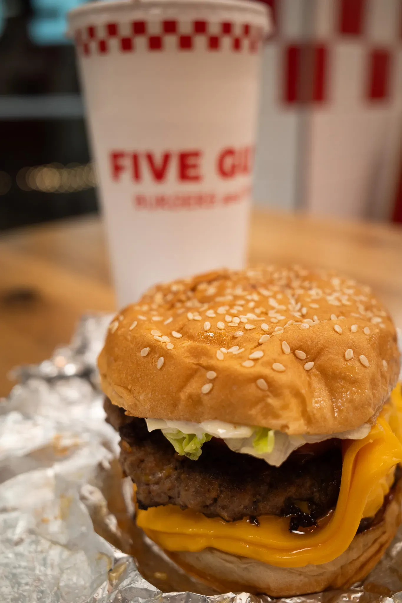 Five Guys cheeseburger with sesame bun, lettuce and melted cheese on foil, a branded red and white cup behind it