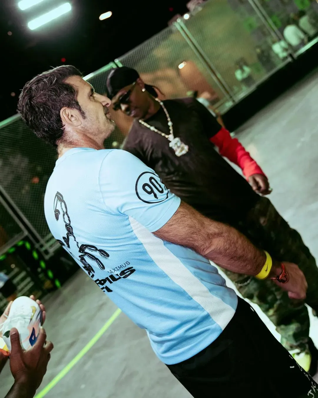 Player in a light blue Scorpius FC jersey greets a man in durag and sunglasses at the edge of the caged pitch