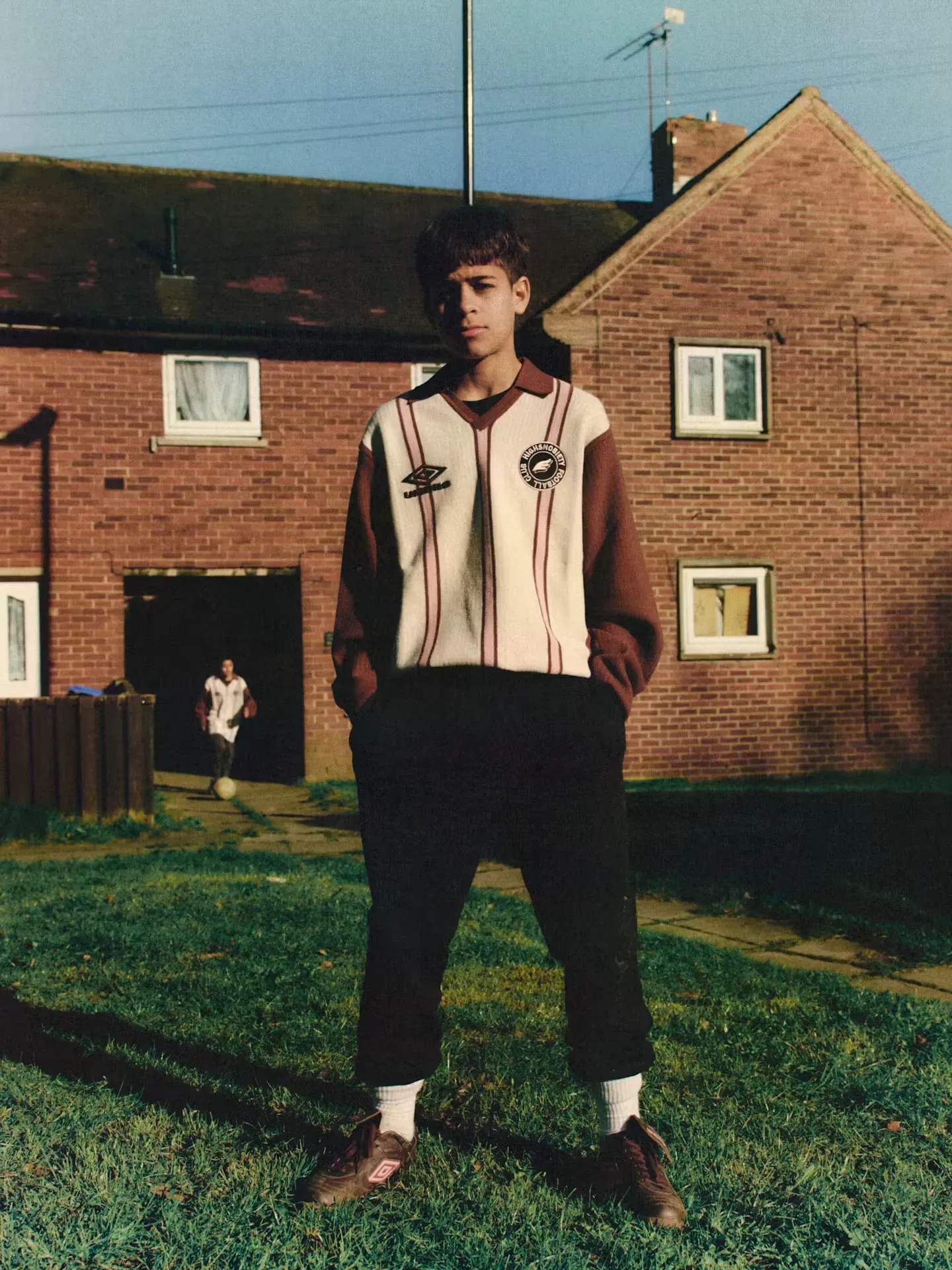 Teenage boy in a striped Umbro and Highsnobiety Football Club knit shirt and brown Umbro boots on grass by brick houses