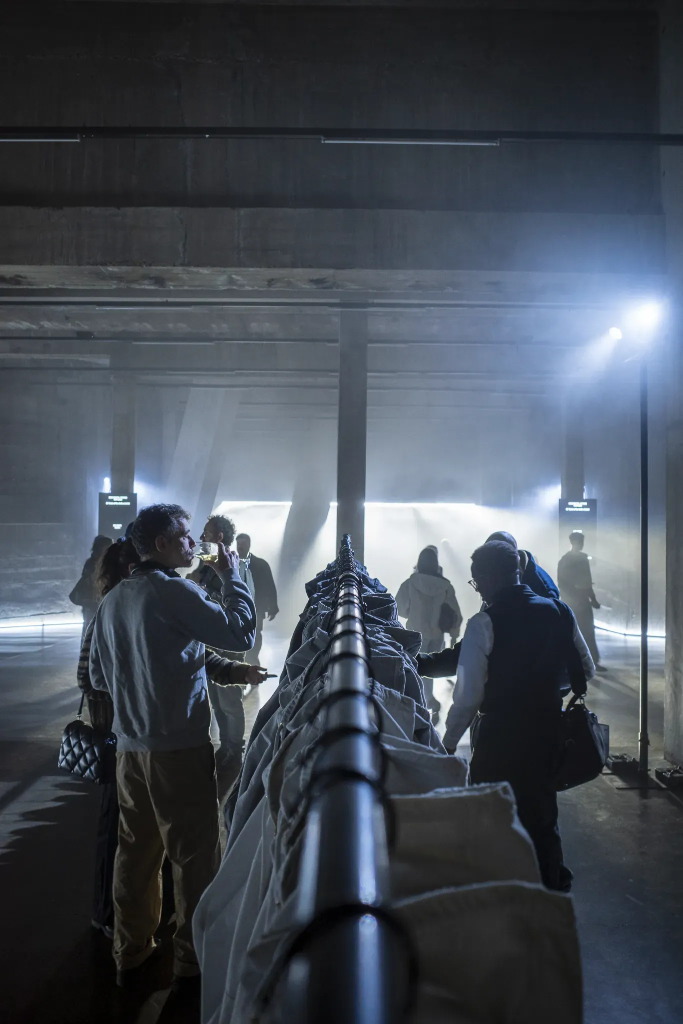 Guests drink and browse along a long rail of pale jackets in a hazy concrete hall lit by bright spotlights
