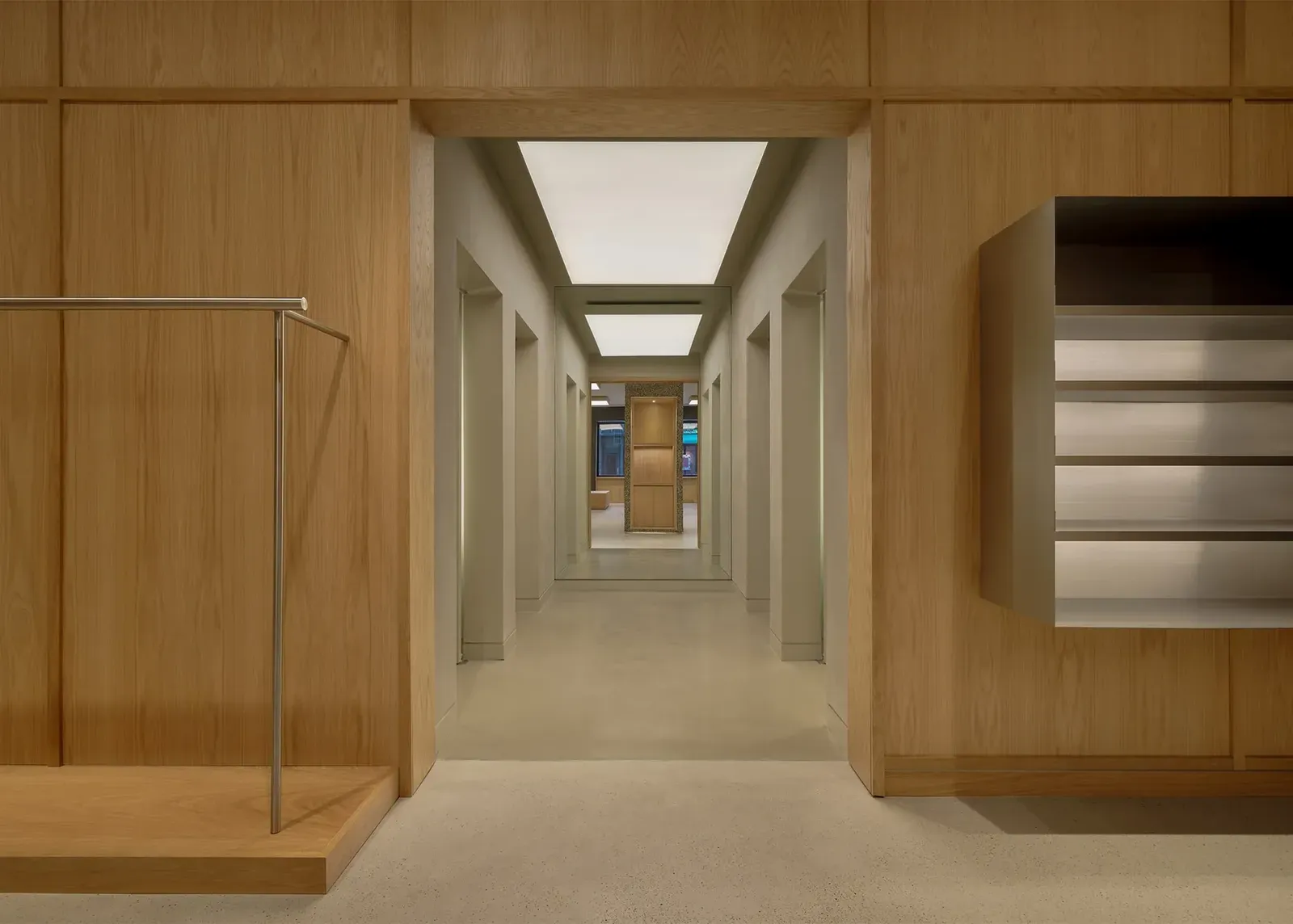Doorway in an oak-panelled wall, flanked by a steel rail and a shelf box, opening onto a pale corridor of fitting rooms