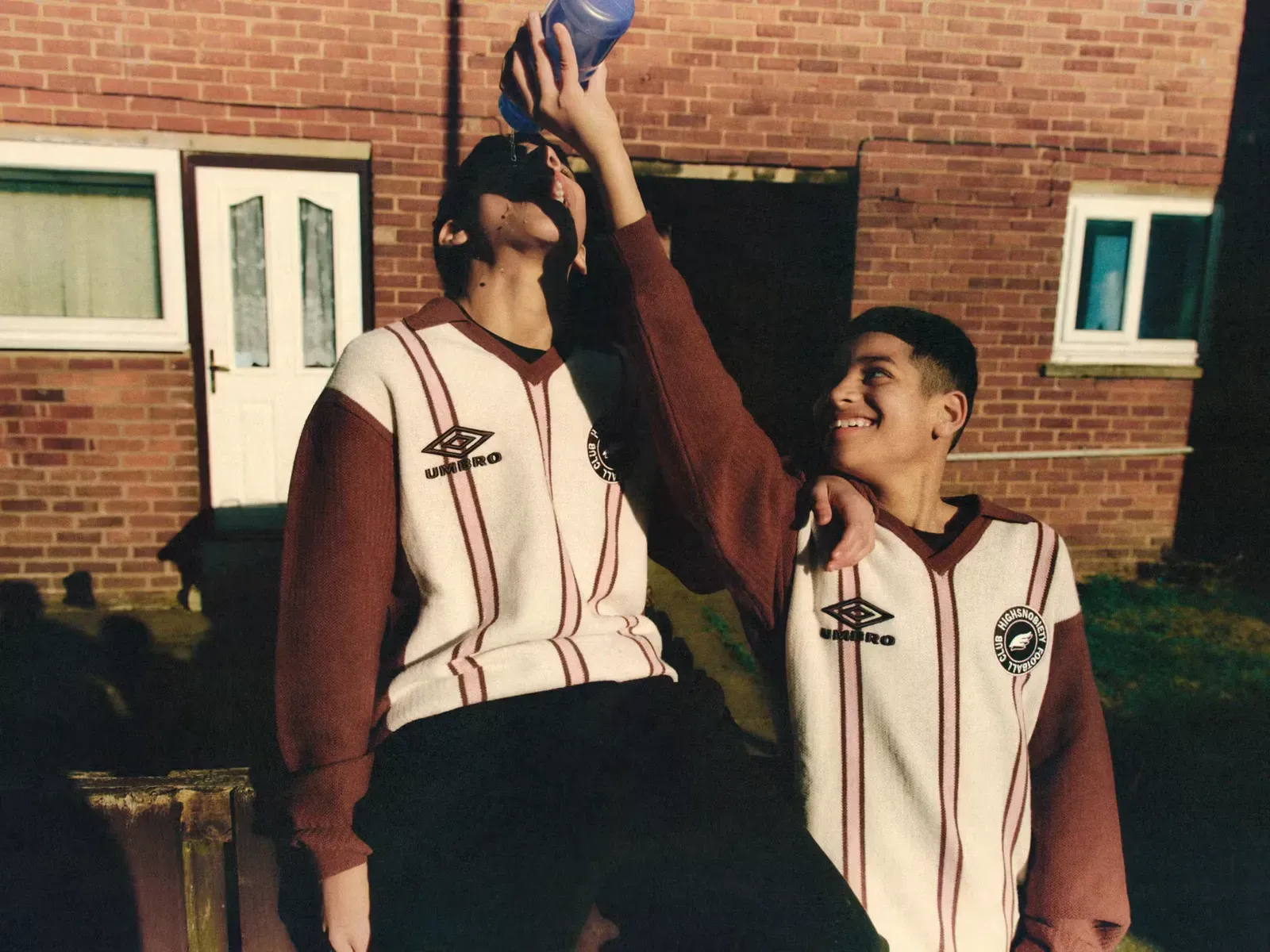 Two boys in matching cream and maroon Umbro knit shirts, one pouring water from a bottle into his mouth as the other grins