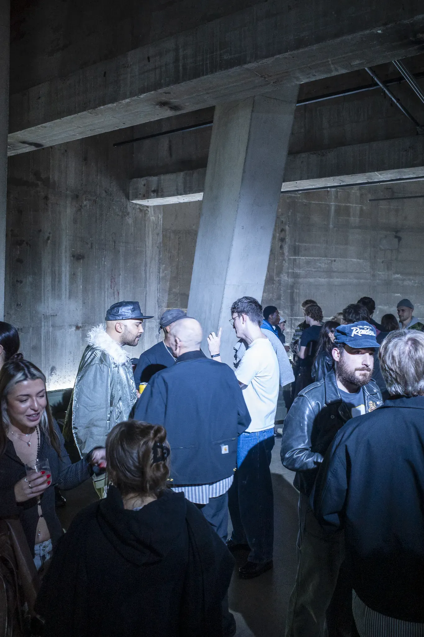 Guests in caps and jackets, one in a dark Carhartt jacket, chat beneath a slanted concrete column