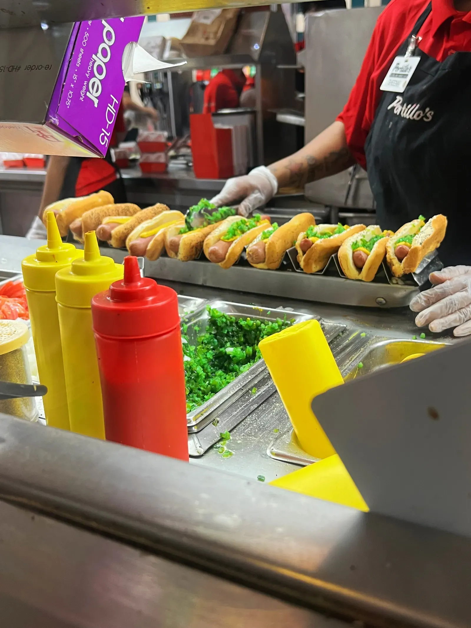 Portillo’s worker in gloves spooning bright green relish onto a row of hot dogs beside mustard and ketchup bottles