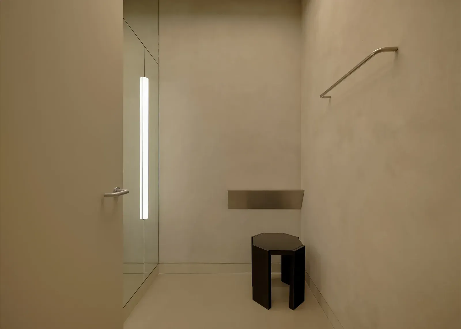 Inside a beige fitting room: mirror with a vertical light tube, black stool, small steel shelf and a bent steel rail
