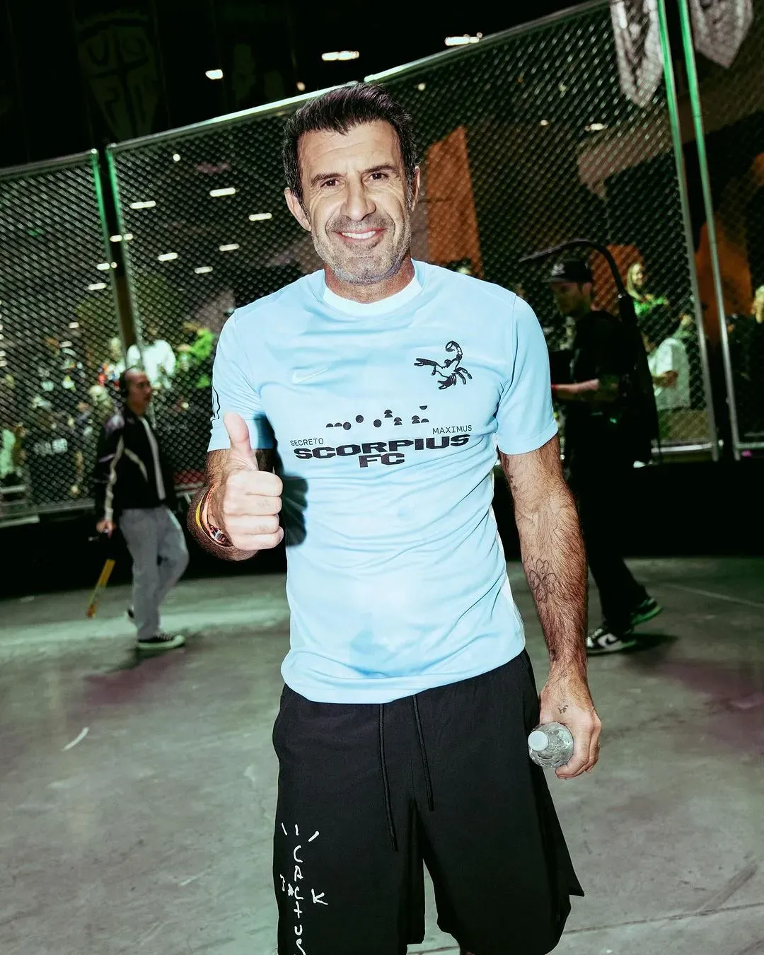 Smiling player in a light blue Nike Scorpius FC jersey gives a thumbs-up, water bottle in hand, outside the cage