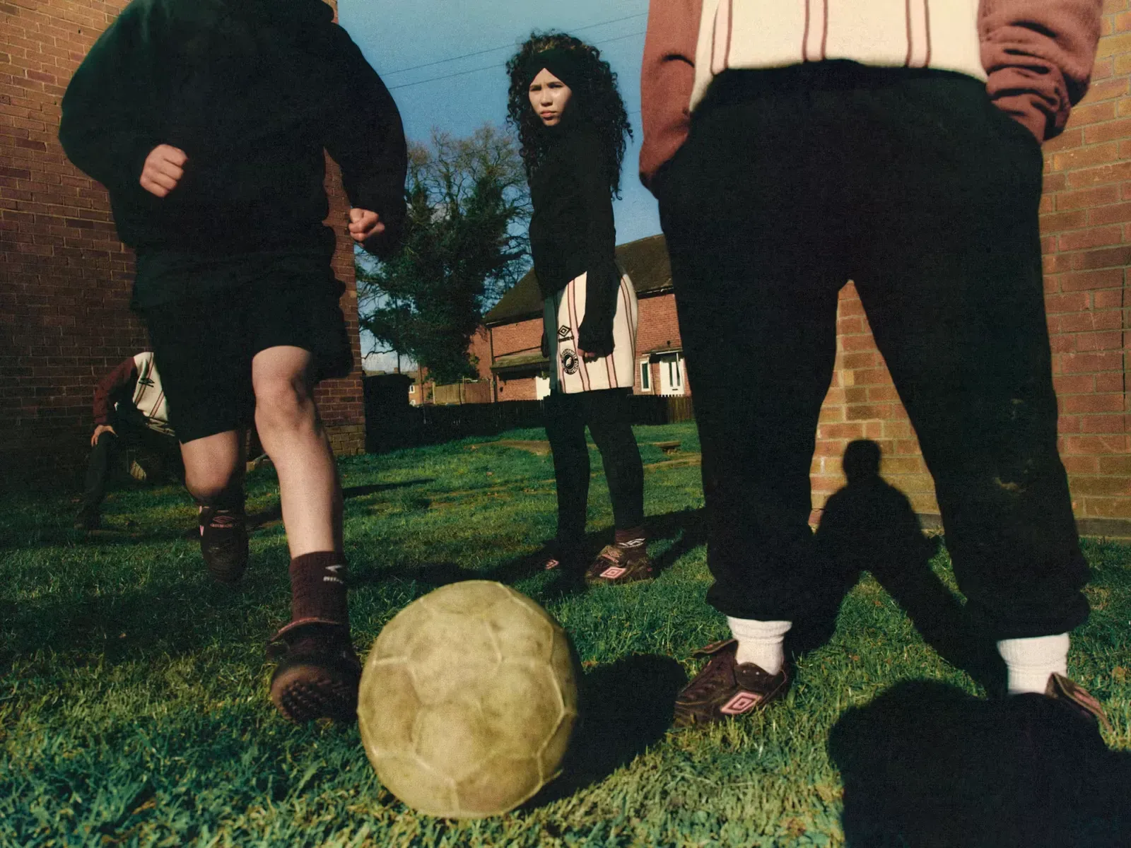 Low-angle view of kids kicking a worn football on grass between brick houses, wearing Umbro boots with pink logos