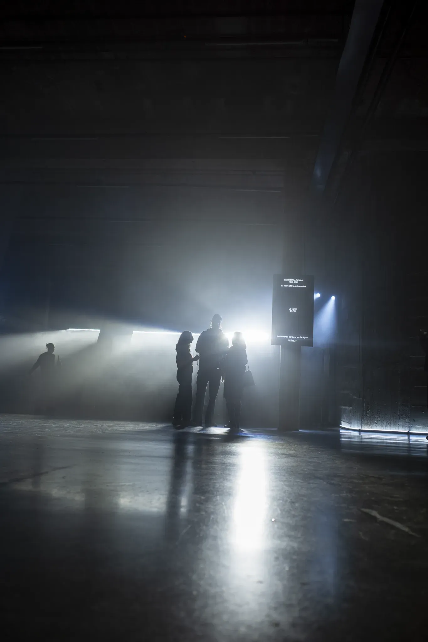 Silhouetted guests by a Sedimental Works sign reading Up next: Klein, in a dark hall filled with haze and light