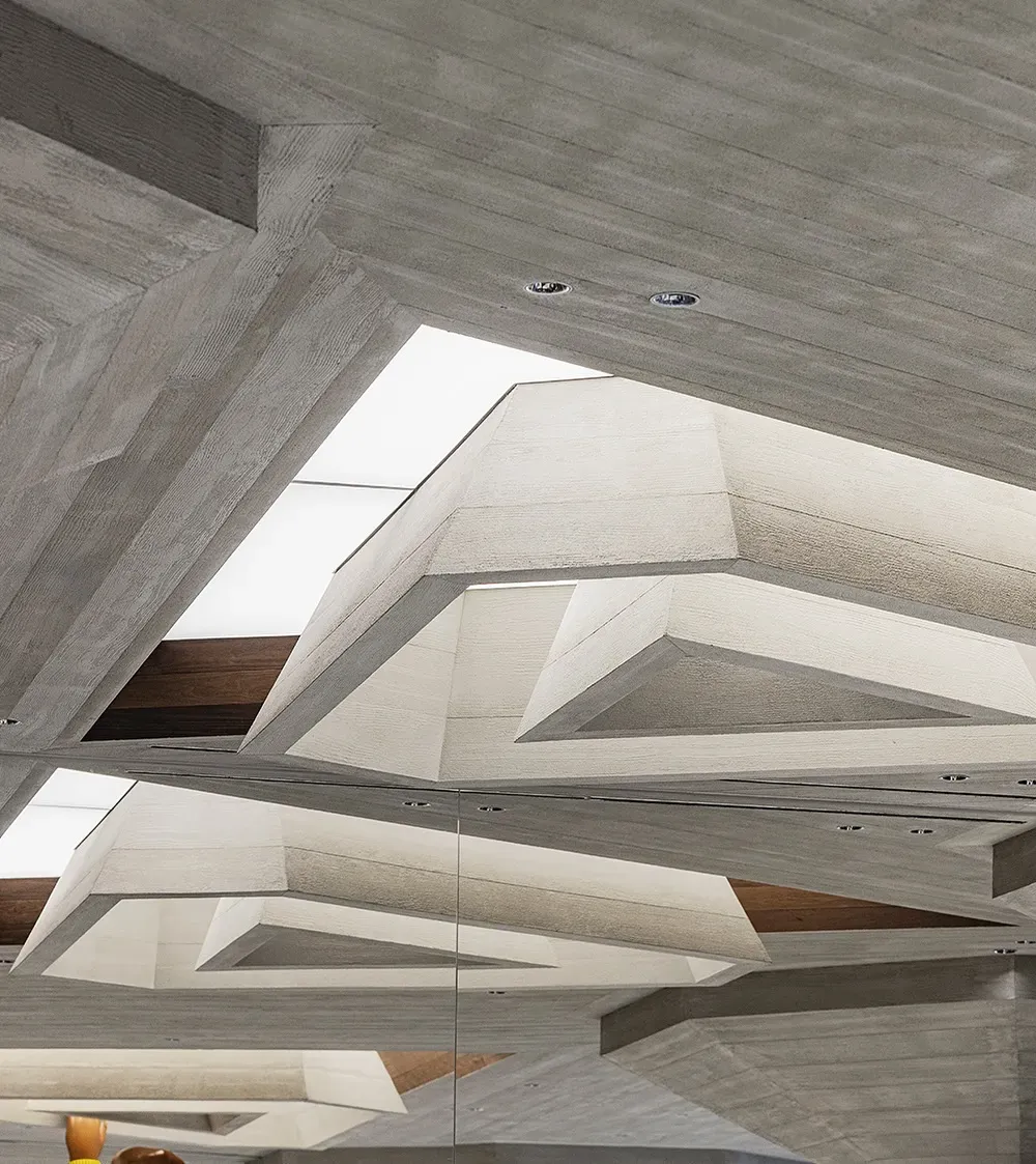 Close-up of the angular board-marked concrete ceiling around a glowing skylight, reflected in a mirrored wall
