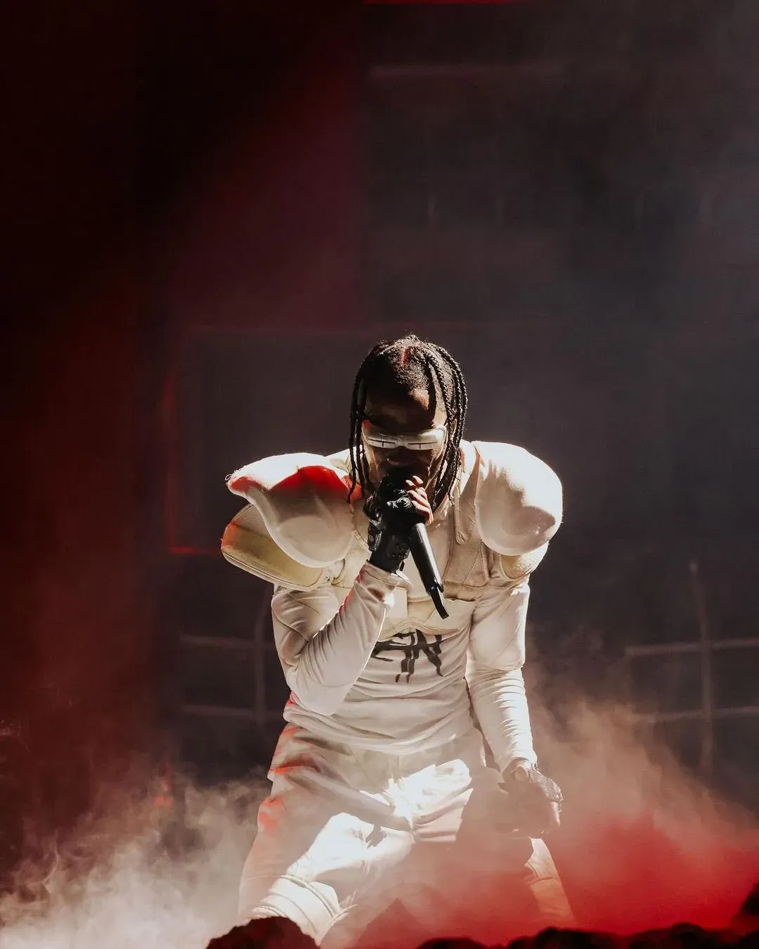 Travis Scott on stage in a white padded outfit and visor, rapping into a microphone through red-lit smoke